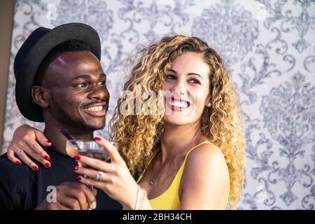 Blonde fille avec cheveux cousue, une robe jaune et des lèvres peintes embrassant un homme noir avec un chapeau tout en buvant un verre de cocktail dans une chambre avec le déc vintage Banque D'Images