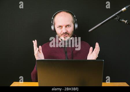 Guy dans hoodie assis derrière le bureau avec ordinateur portable et casque sur fond noir, parler au microphone, enregistrer vidéo blog Banque D'Images