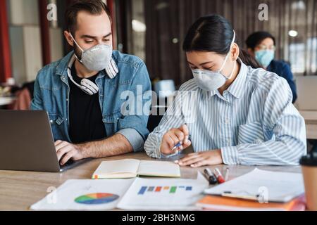 Photo de jeunes étudiants multinationaux dans des masques médicaux étudiant avec ordinateur portable en classe Banque D'Images