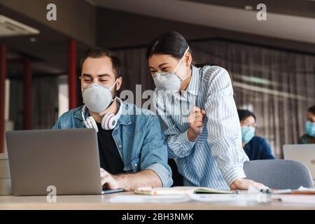 Photo d'étudiants multinationaux joyeux dans les masques médicaux étudiant avec ordinateur portable en classe Banque D'Images