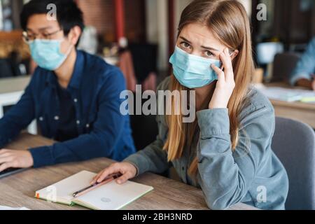 Photo de jeunes étudiants multinationaux avec maux de tête dans les masques médicaux étudiant en classe Banque D'Images