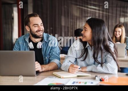 Photo d'étudiants multinationaux joyeux utilisant un ordinateur portable et écrire dans un livre d'exercice pendant qu'ils étudient en classe Banque D'Images