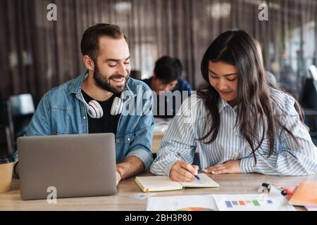 Photo d'étudiants multinationaux joyeux utilisant un ordinateur portable et écrire dans un livre d'exercice pendant qu'ils étudient en classe Banque D'Images