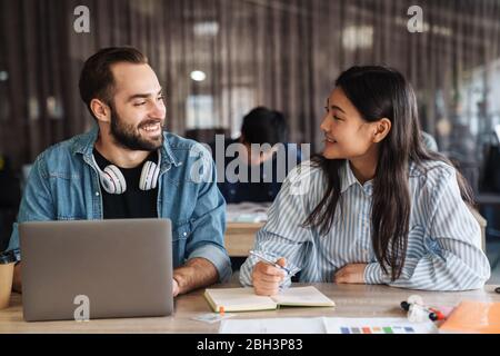 Photo d'étudiants multinationaux joyeux utilisant un ordinateur portable et écrire dans un livre d'exercice pendant qu'ils étudient en classe Banque D'Images