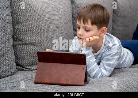 Formation à distance en ligne. Enfant d'école en auto-isolation en utilisant une tablette pour les devoirs, ennuyé enfant triste visage allongé tête vers le bas regardant profondément dedans Banque D'Images