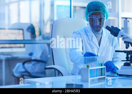 Jeune médecin africain dans des lunettes de protection travaillant avec des échantillons tout en étant assis à son lieu de travail devant le microscope Banque D'Images