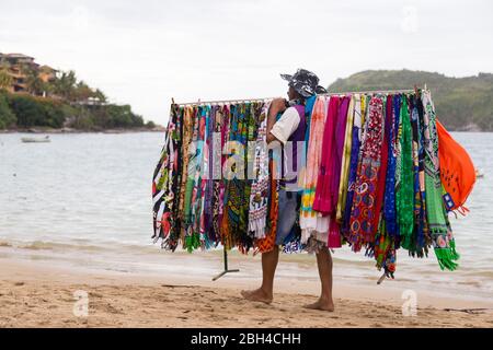 Vendeur de plage typique brésilienne marchant sur la plage et portant pour la vente des robes de plusieurs couleurs. FERRADURA BEACH, BUZIOS, BRÉSIL. Banque D'Images