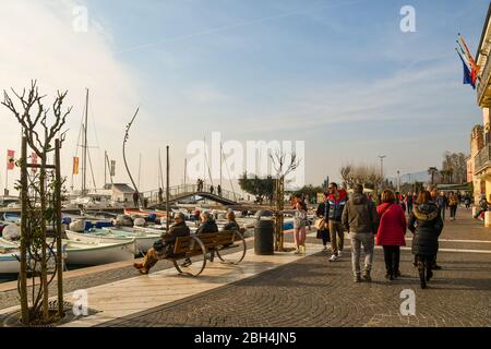 Vue sur le lac bondé avec le port et les gens marchant et assis sur des bancs dans une journée ensoleillée d'hiver, Bardolino, Vérone, Vénétie, Italie Banque D'Images