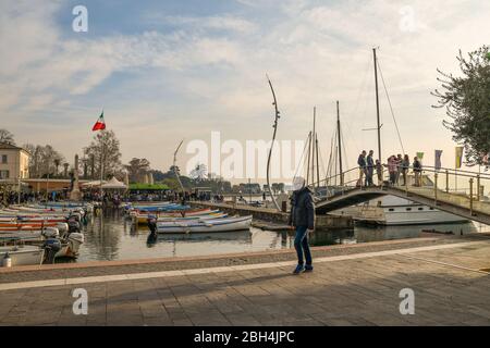 Port de la vieille ville sur la rive du lac de Garde avec bateaux amarrés, un pont piéton et des gens à un festival en arrière-plan, Bardolino, Italie Banque D'Images