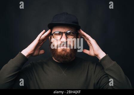 Concentré. Ma tête fait mal. J’ai mal à la tête. Homme barbu avec les yeux fermés en touchant la tête sur fond noir. Banque D'Images