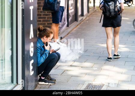 Londres, Royaume-Uni - 26 juin 2018 : une femme assise sur la rue du trottoir à Covent Garden centre ville vaporing avec vapeur d'ecigarette Banque D'Images