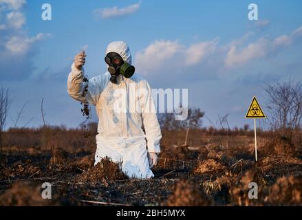 Homme scientifique, portant un costume de protection blanc, un masque à gaz et des gants, debout sur les genoux entre les anthills et déversant le sol de sa main, signe de biodanger derrière lui. Concept d'écologie, terre brûlée Banque D'Images