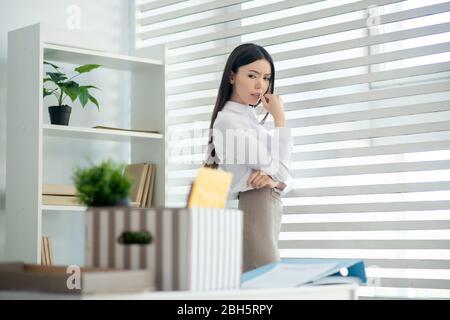 À la maison. Jeune brunette femme debout à la fenêtre, regardant sa boîte avec des trucs personnels, debout sur le bureau Banque D'Images