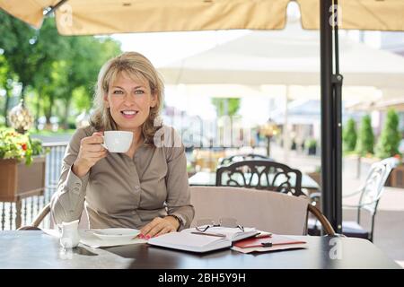 Portrait de belle femme blonde mûre regardant latéralement, espace de copie. Femme d'affaires souriante d'âge moyen, en train de boire du café dans un café extérieur Banque D'Images