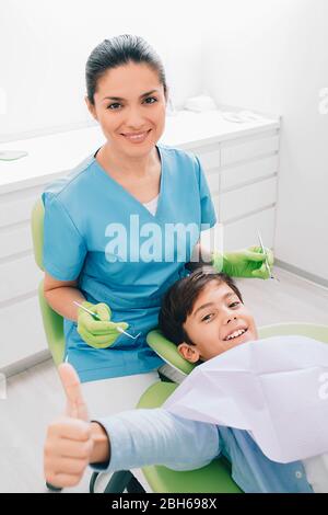 Un petit garçon heureux qui montre des pouces. Il visite le dentiste pédiatre Banque D'Images