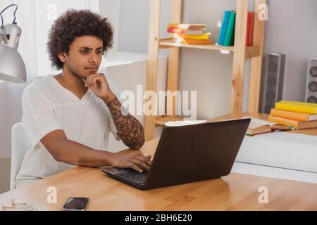 Homme pensif, avec des cheveux raillés et vêtus de manière informelle, regardant attentivement l'ordinateur portable tout en étant assis à son lieu de travail à la maison Banque D'Images