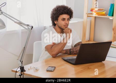 Homme pensif, avec des cheveux raillés et vêtus de manière informelle, regardant attentivement l'ordinateur portable tout en étant assis à son lieu de travail au bureau Banque D'Images