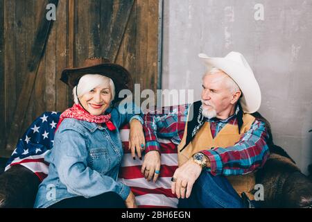 Couple âgé vêtu de cowboy, assis sur un canapé avec un drapeau américain. Célébrer le jour de l'indépendance de l'Amérique Banque D'Images