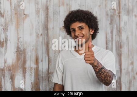 L'homme aux cheveux bouclés positif avec de belles tattoo showing Thumbs up. Tout en se tenant à l'ancien fond de bois Banque D'Images