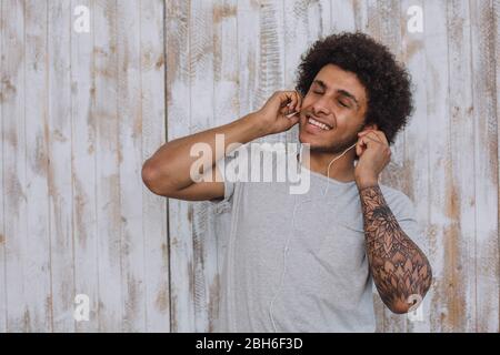 Profitez du son. Homme mulâtre, avec les cheveux bouclés portant des écouteurs, ferma les yeux et souriant contre mur en bois rétro Banque D'Images