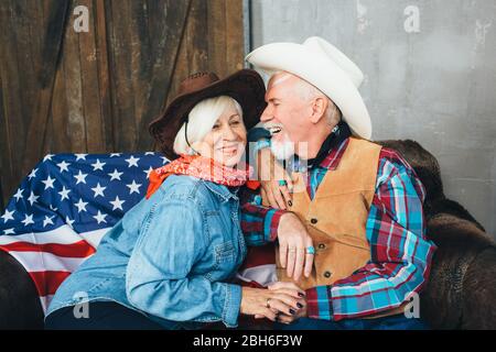 couple âgé, vêtu de chapeaux de cowboy, rire, se prenant les mains de l'autre. Derrière, sur le canapé se trouve le drapeau américain, la célébration de l'Amérique dans Banque D'Images