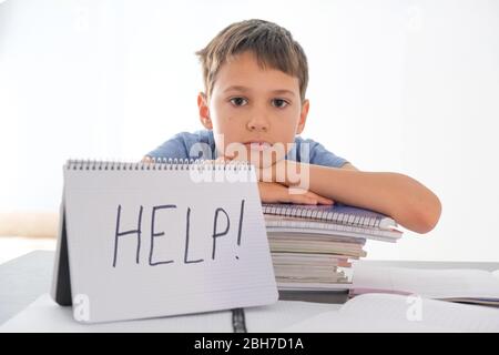 Un garçon fatigué assis à la table avec de nombreux livres. L'aide Word est écrite sur le bloc-notes. Difficultés d'apprentissage, éducation, apprentissage en ligne à la maison Banque D'Images