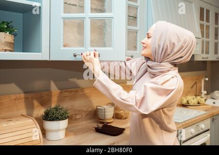 Jolie femme de ménage musulmane dans le hijab ouvre une armoire de cuisine sur la cuisine moderne vue latérale Banque D'Images