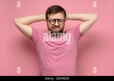 L'homme barbu avec des lunettes n'est pas dans l'esprit, il grimaces en exaspération, mettant les mains derrière la tête, sur un fond rose. Banque D'Images