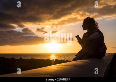 belle femme en silhouette lors d'un magnifique coucher de soleil sur l'océan à l'aide d'un smartphone mobile technolofy assis sur un long beanch en pierre. de bonnes vacances en vacances avec des amis Banque D'Images