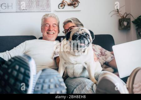 Un couple caucasien s'assoit sur le canapé avec un chien de pug drôle regardant. Bonheur avec amour pour les animaux et le meilleur concept d'ami - la vie quotidienne à la maison avec les gens agréables et le vieux chiot Banque D'Images