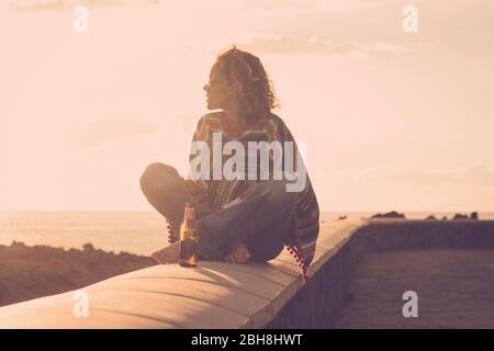 Jolie femme indépendante et forte assise sur un mur pieds nus et style hippy regardant la nature autour et profiter d'un coucher de soleil doré extérieur de style de vie de loisirs près de l'océan - style de vie coloré Banque D'Images