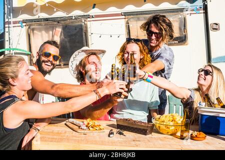 Groupe d'amis heureux à craquer et à clinquer ensemble la bière avec le bonheur - les gens agréables avec la nature et le style de vie extérieur - les garçons et les filles souriant et riant avec une caravane alternative en arrière-plan Banque D'Images