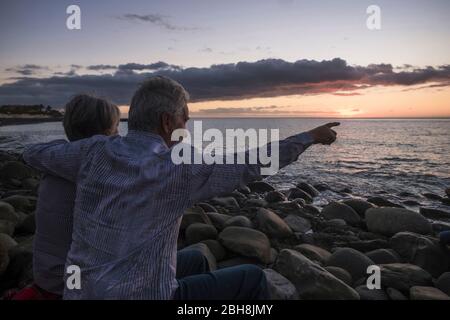 Romance pour couple senior adulte assis à la plage à la recherche d'un coucher de soleil coloré sur la mer - vacances et concept d'amour pour homme mûr et femme dans l'amour - scène romantique Banque D'Images