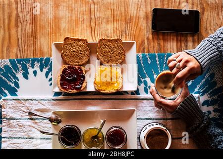Point de vue vertical de la femme qui fait le petit déjeuner à l'hôtel ou à la maison - pain et mermalades et café temps pour la nourriture saine d'énergie pour commencer la journée - téléphone portable noir sur la table en bois Banque D'Images