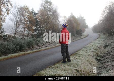 Jeune homme dans une veste rouge en duvet marchant le long d'une route écossaise à Rosehall, Lairg, Scotland Highlands, Royaume-Uni Banque D'Images