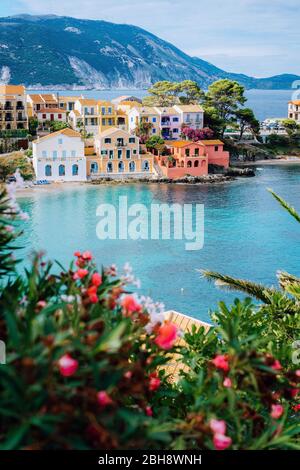 Belles maisons colorées du sud et baie bleue de la mer Méditerranée avec quelques fleurs rouges au premier plan. Village d'Assos à Kefalonia, Grèce. Prise de vue verticale. Banque D'Images