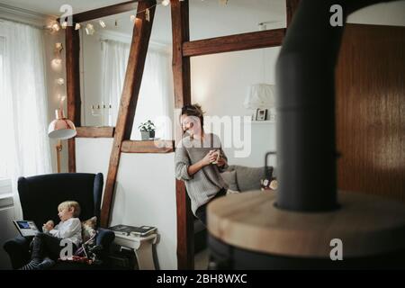 junge Frau in ihrer Wohnung, hält Tasse in der Hand, lächelen, Sohn spielt mit Tablet Computer Banque D'Images