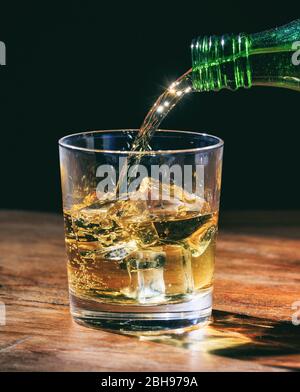 Bouteille verte qui verse du whisky sur les rochers, en verre cristal sur table en bois. Portrait vertical de la boisson alcoolisée avec des glaçons sur fond sombre. Banque D'Images