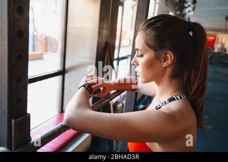 Jeune sportif séduisant debout dans la salle de gym, se reposant après des exercices, se penchant sur un rail, en vérifiant la montre de fitness Banque D'Images