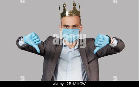 Jeune homme d'affaires dans une couronne et un masque de protection et des gants. Rester à la maison pendant la quarantaine du coronavirus Covid-19. Montre pouce vers le bas Banque D'Images