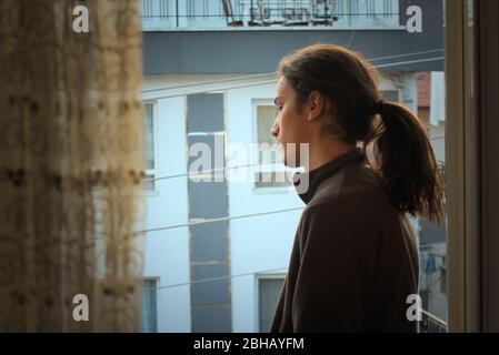 Adolescent millénaire isolé et déprimé avec de longs cheveux regardant les rues vides de son balcon avec tristesse Banque D'Images