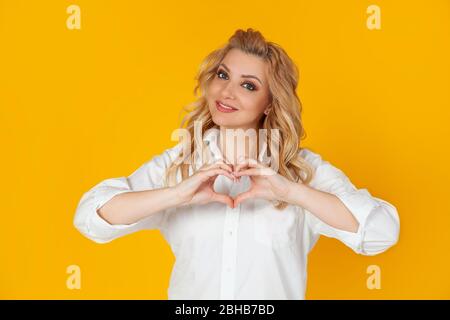 Charmante femme blonde romantique et charismatique passionnée d'yeux bleus montre coeur geste de reconnaissance sympathie look sympathique appareil photo souriant, femme exprimer de vrais sentiments. Aimez tout ce dont vous avez besoin. Banque D'Images