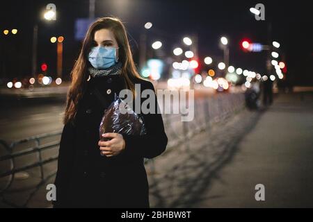 Portrait de la jeune femme triste portant un masque médical portant du pain tout en marchant dans la rue de la ville la nuit Banque D'Images