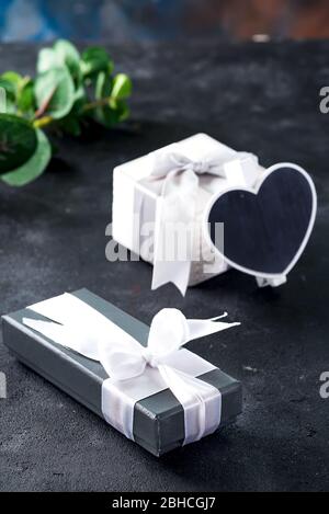 Carte festive avec deux boîtes cadeaux attachées de rubans blancs, tableau de surveillance en forme de coeur et branche de plantes naturelles vertes sur une pierre noire b Banque D'Images