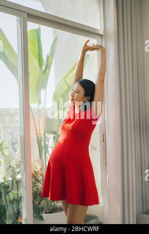 Portrait de la jeune femme enceinte s'étirant joyeusement debout dans la salle de séjour éclairée par la lumière du soleil chaude, espace de copie Banque D'Images