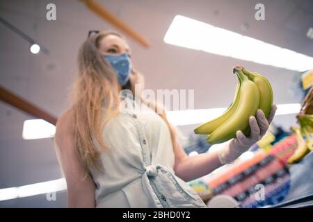 Une jeune femme d'une épicerie portant un masque facial et des gants jetables se trouvant dans le département de fruits de l'épicerie. Les personnes portant des masques en tissu Banque D'Images