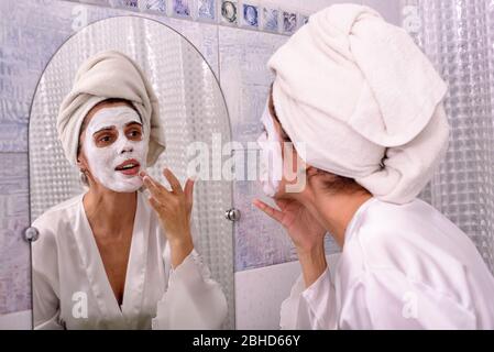 Belle femme brunette à la maison dans la pyjama et serviette sur sa tête mettre le masque blanc sur son visage à la salle de bains Banque D'Images