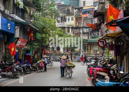 Hanoi, Vietnam, 30 décembre 2019 - vue arrière sur une vieille dame, vendeur de nourriture de rue, poussant son vélo plein de fruits à vendre, au milieu d'une rue Banque D'Images