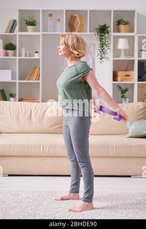 Blonde active femme dans le t-shirt et les leggins debout sur le sol du salon et utilisant le groupe flexible tout en exerçant dans l'environnement domestique Banque D'Images