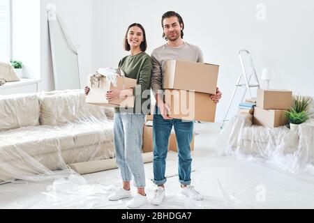 Jeune couple joyeux en vêtements de travail tenant des boîtes emballées avec des affaires ménagères tout en se tenant dans la salle de séjour de nouveau plat Banque D'Images